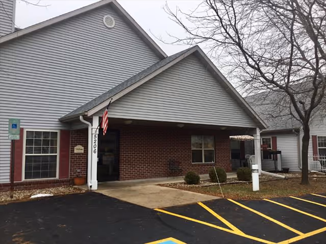 Exterior view of McFarland Villa Assisted Living building entrance with a covered porch, an American flag hanging near the door, a handicap parking sign, and leafless trees in the surrounding area.