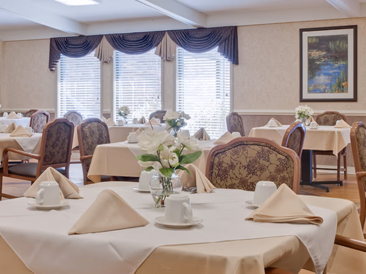 Dining room with tables set with folded napkins, cups, and floral centerpieces and upholstered chairs.