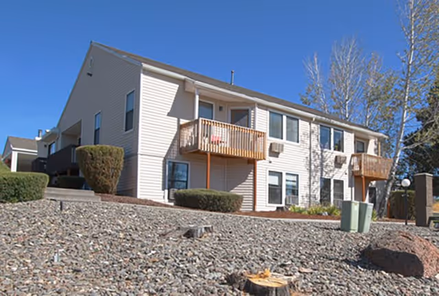 Exterior view of a two-story residential building with beige siding, small balconies, and several windows. The foreground shows a rocky landscape with trimmed bushes and a clear blue sky above.