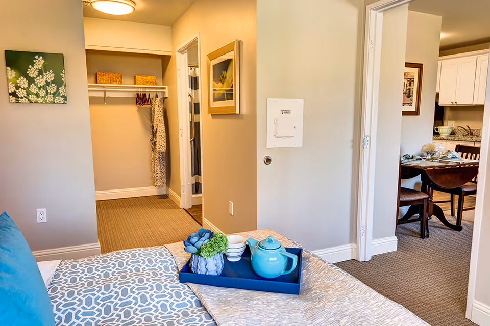 View from a bedroom showing a bed with a blue pillow and a tray holding a blue teapot, cups, and a small plant. The room opens into a hallway with a closet containing hanging clothes and baskets on a shelf. To the right, there is a dining area with a table set for a meal and a kitchen with white cabinets and granite countertops.