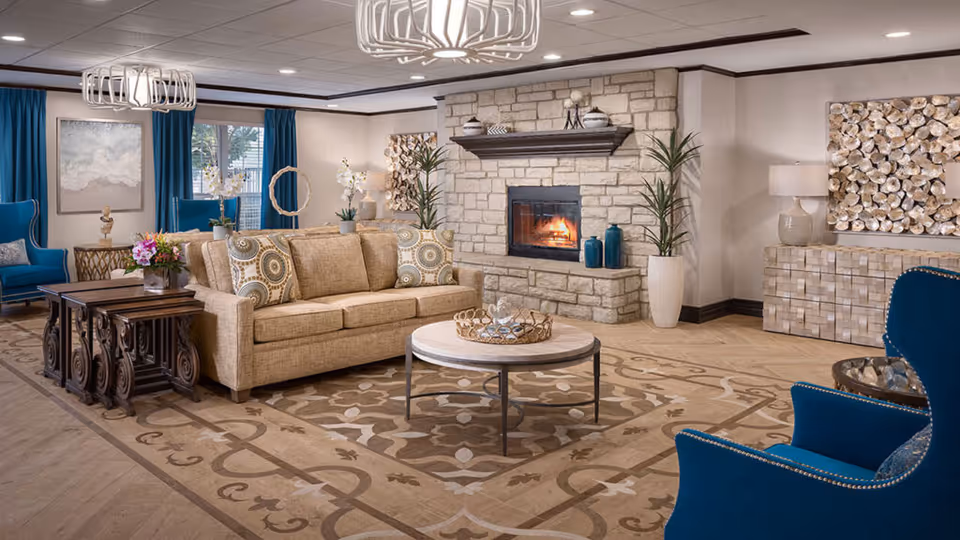 A cozy senior living facility common area with a beige sofa adorned with patterned cushions, a round coffee table, and two blue armchairs. The room features a stone fireplace with a fire burning, decorative plants, and artwork on the walls. The floor has a patterned rug, and the windows are dressed with blue curtains.
