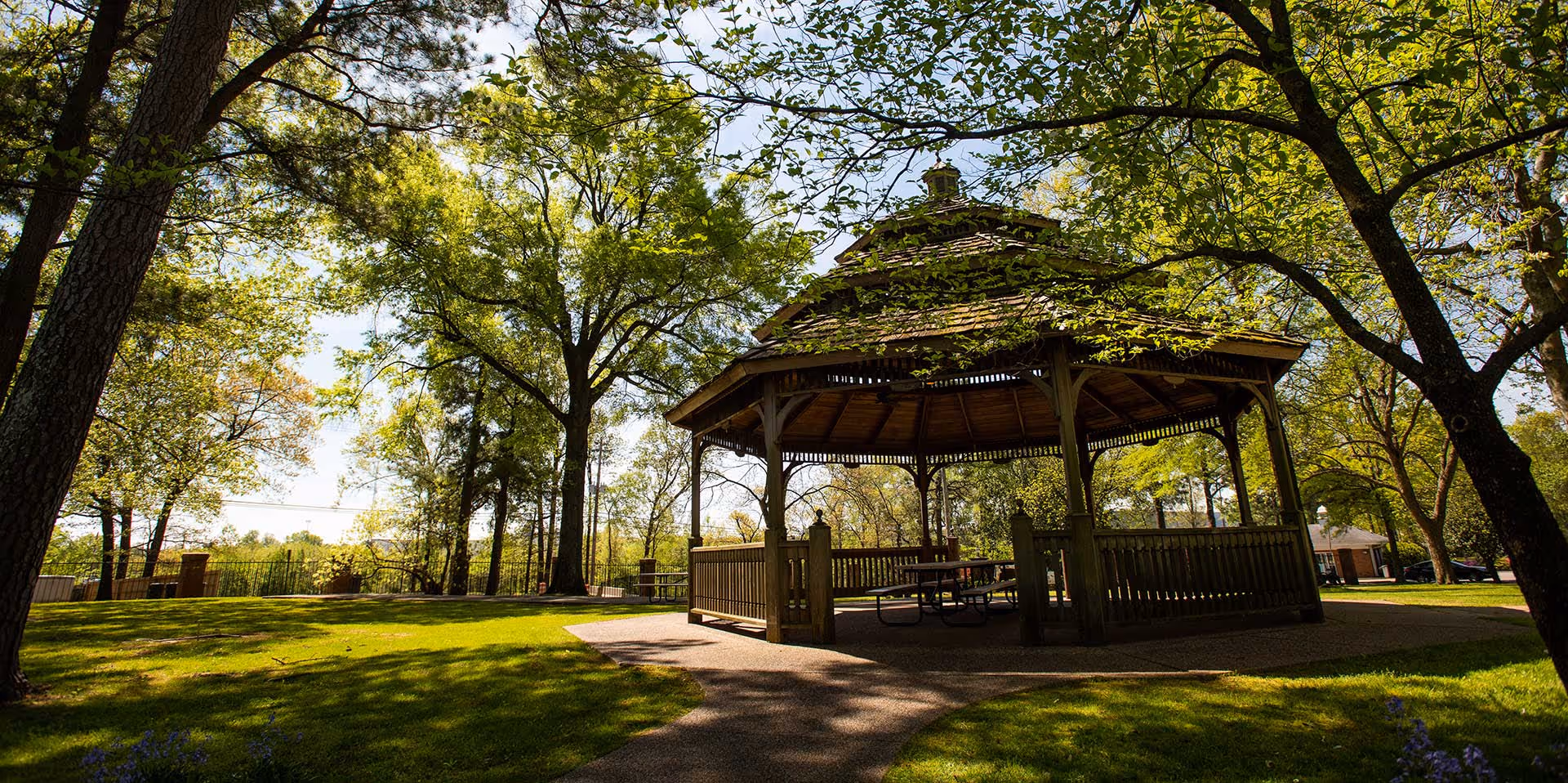 A wooden gazebo with a shingled roof situated in a green park-like area with trees and grass, under a sunny sky.