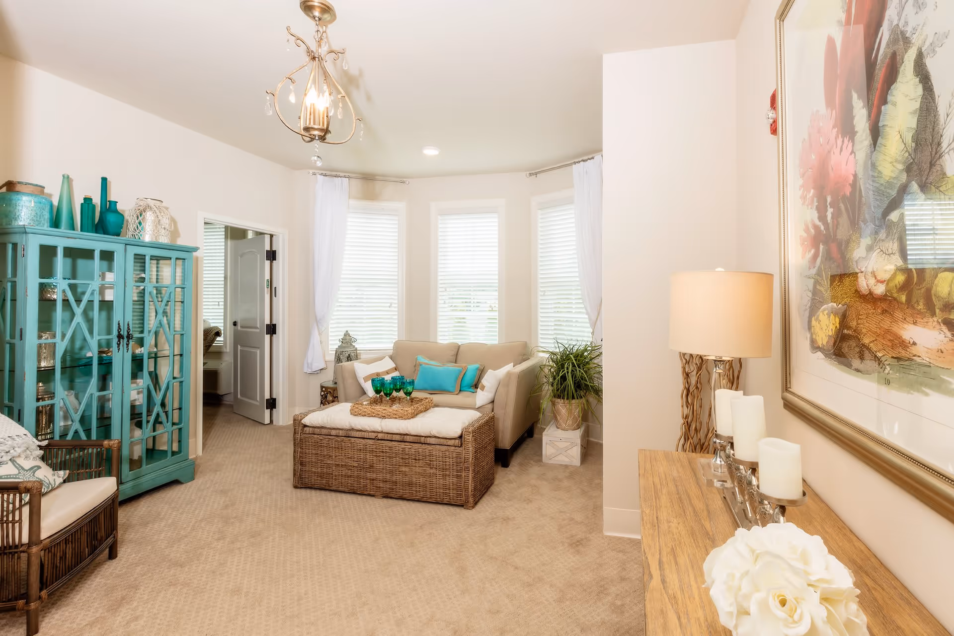 A cozy living room with beige carpet and walls, featuring a beige sofa with turquoise and white pillows, a wicker ottoman with a tray holding green glasses, a turquoise cabinet with decorative items, a wicker chair with a cushion, a wooden console table with candles and a lamp, and a large framed floral artwork on the wall. Three windows with white blinds and sheer white curtains allow natural light into the room.