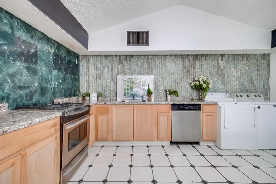 A kitchen area with light wood cabinets, a stainless steel oven and dishwasher, a white washing machine and dryer, granite countertops, green marble and tile backsplash, and decorative plants and flowers on the countertop.
