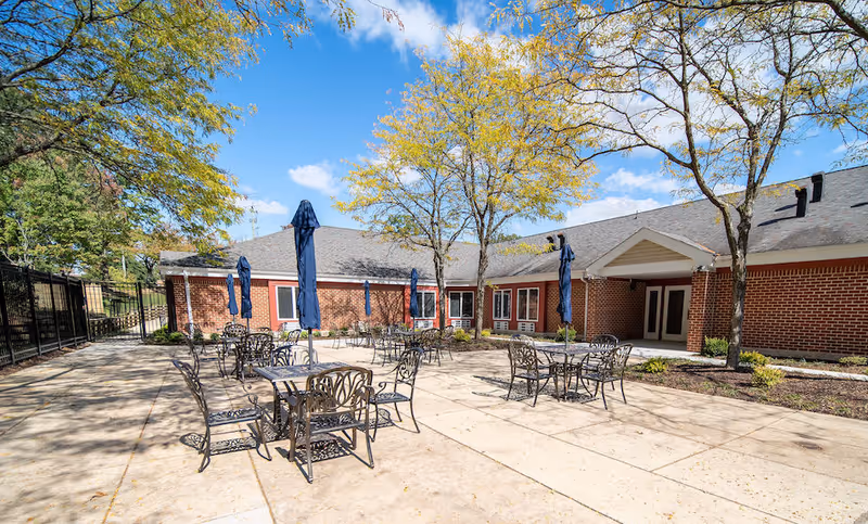 Outdoor patio area at Independence Court of Hyattsville with several metal tables and chairs, each table having a closed blue umbrella. The patio is surrounded by a brick building and some trees with yellowing leaves under a bright blue sky with scattered clouds.