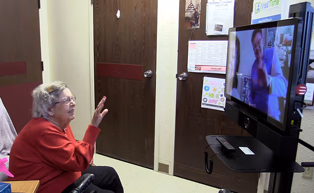 An elderly woman in a wheelchair wearing a red sweater is waving and smiling at a large screen displaying a video call with another person who is also waving.