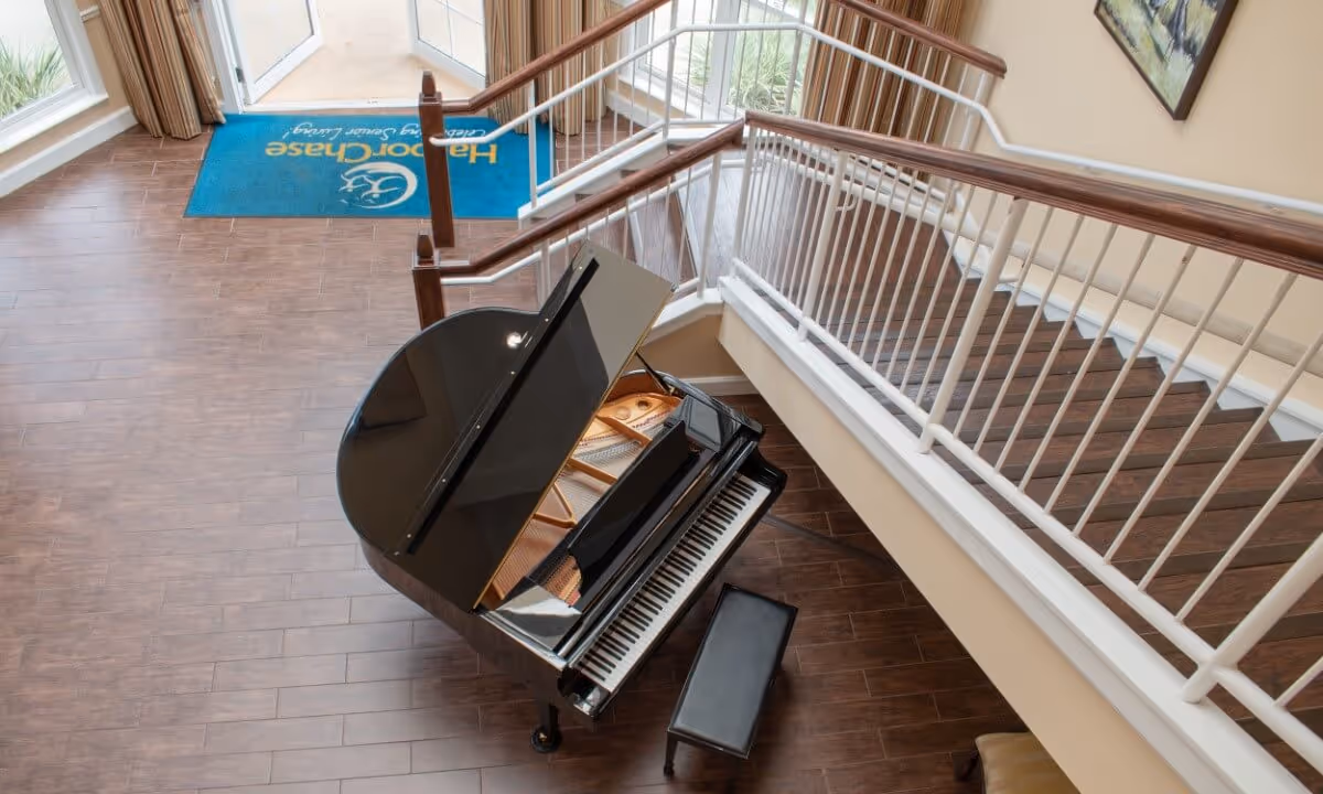 Interior lobby with a black grand piano and bench beneath a staircase and railing by the entryway.