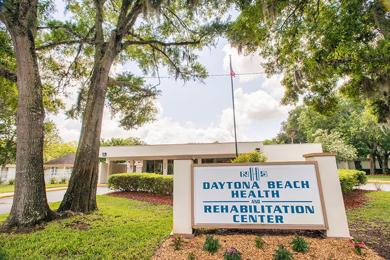 Exterior view of Daytona Beach Health and Rehabilitation Center showing a white building partially obscured by trees and bushes, with a large sign in front displaying the facility's name. The sky is partly cloudy and there is an American flag on a pole behind the sign.