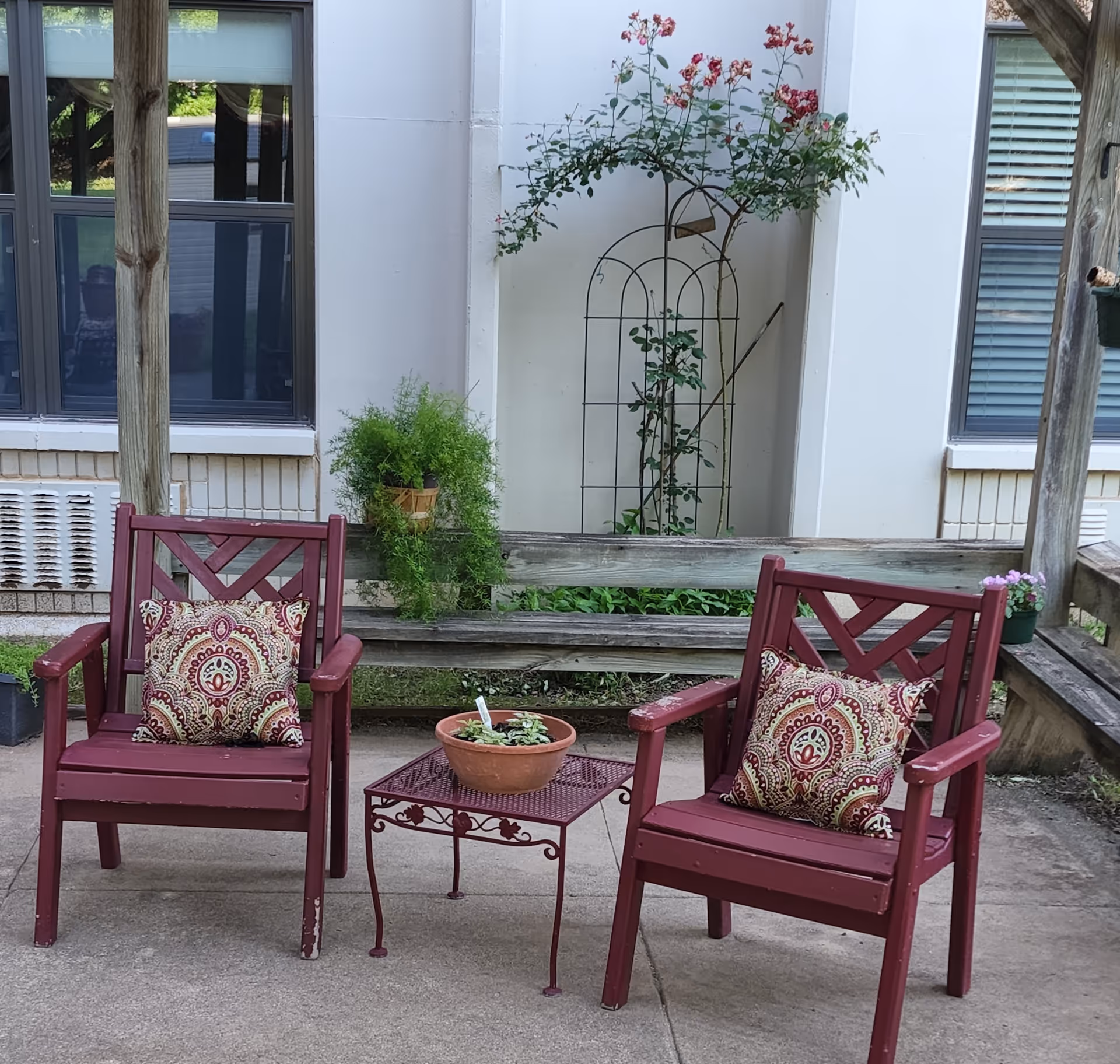 Two red wooden chairs with patterned cushions are placed on a concrete patio. Between them is a small red metal table with a potted plant. Behind the chairs is a wooden bench with a hanging potted plant and a trellis with climbing flowers against a white building wall with windows.