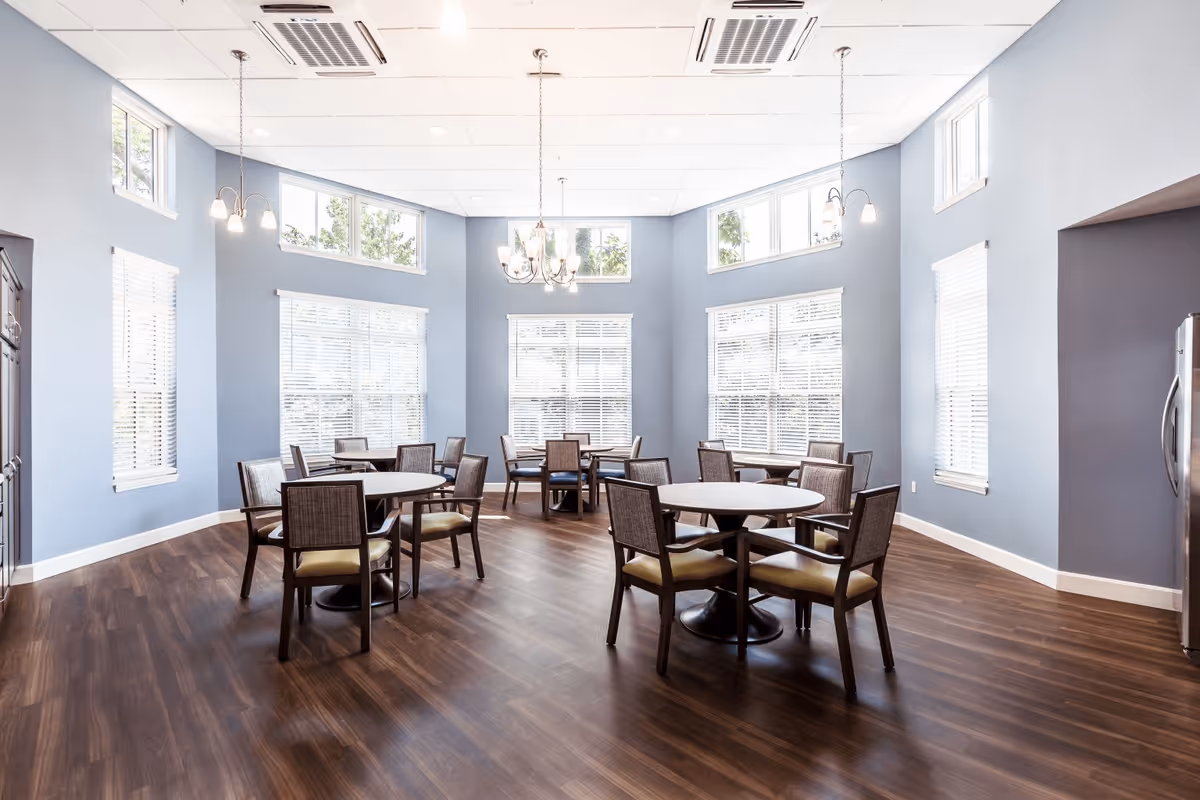 A bright dining room with several round tables and chairs arranged on a wooden floor. The walls are painted light blue and feature multiple large windows with white blinds, allowing natural light to fill the space. There are hanging light fixtures and a chandelier providing additional lighting.