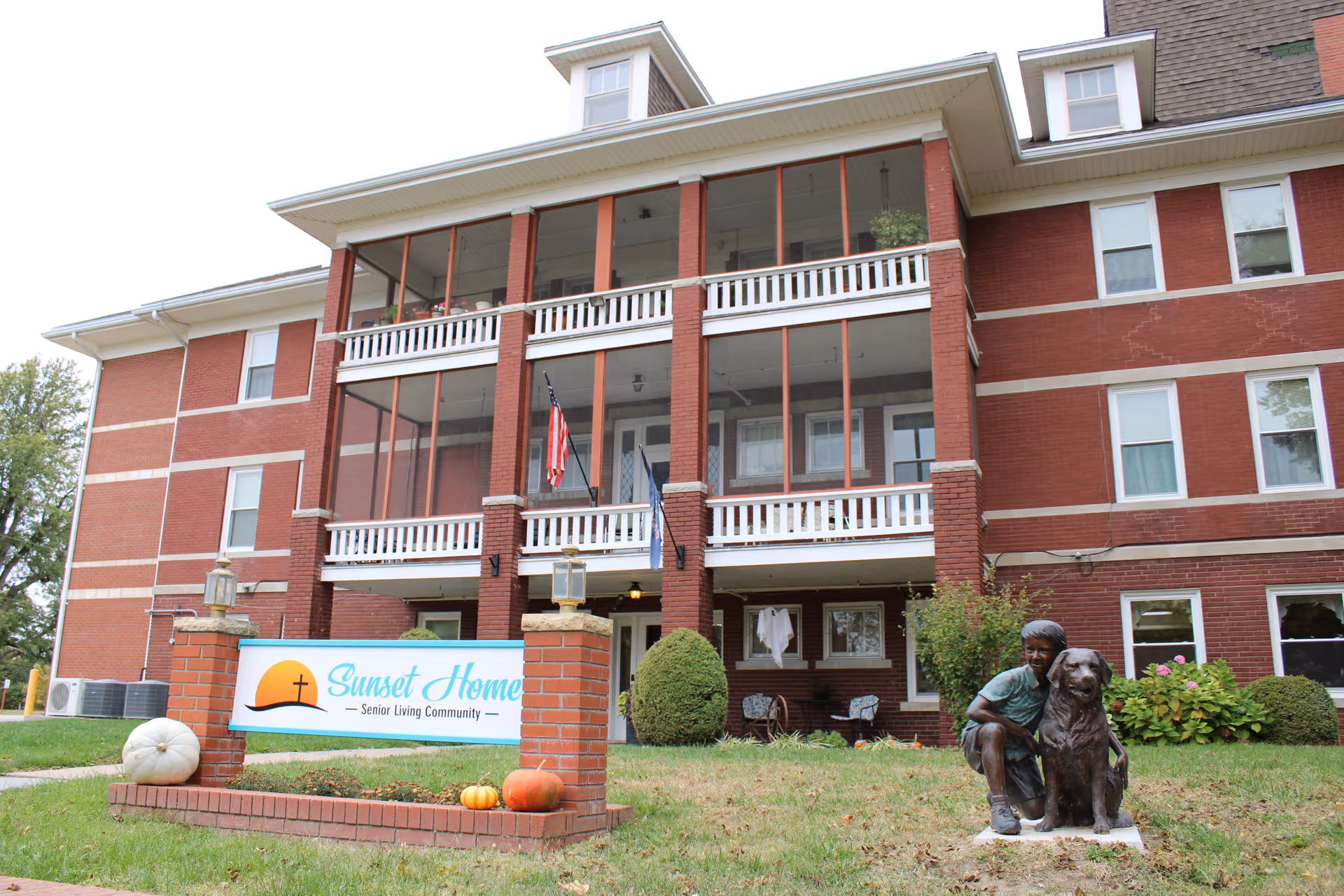 Front exterior view of a three-story red brick senior living community building with screened balconies. In front of the building is a sign that reads 'Sunset Home Senior Living Community' and a statue of a child crouching next to a large dog. There are pumpkins and greenery around the sign and statue.