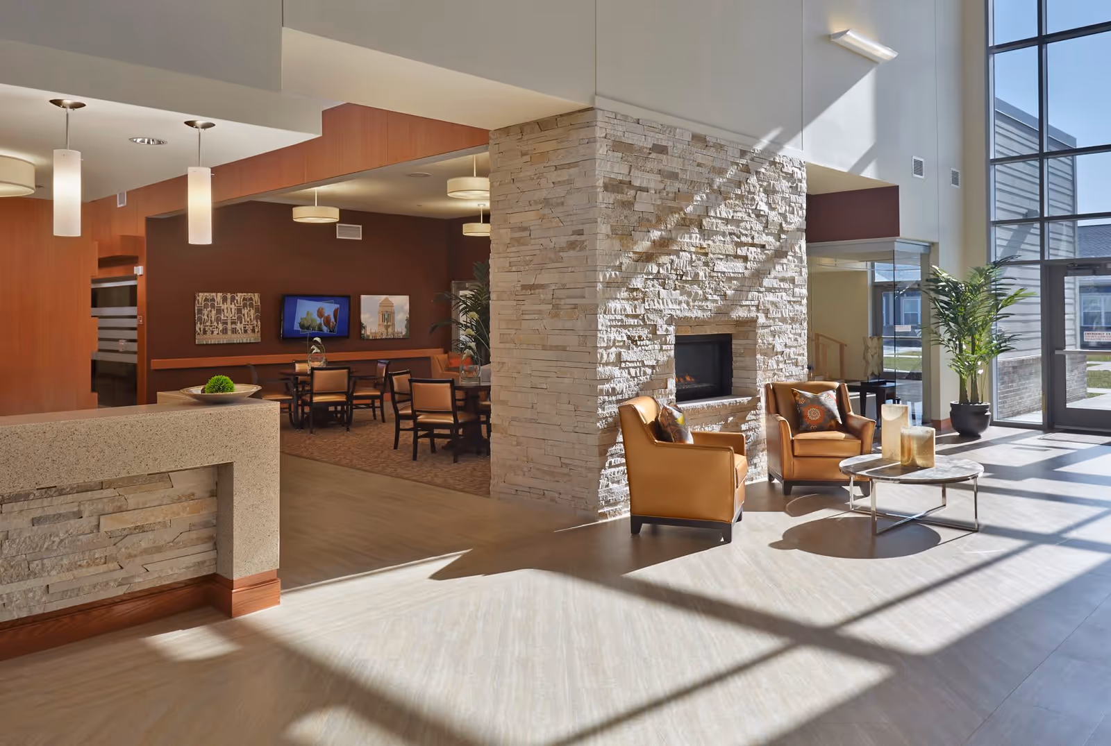 A bright and spacious interior common area with large windows letting in natural light. The room features a stone fireplace with two brown leather armchairs and a round coffee table in front. To the left, there is a dining area with tables and chairs, a TV mounted on a dark brown wall, and decorative artwork. The floor is light-colored, and there are modern pendant lights hanging from the ceiling.
