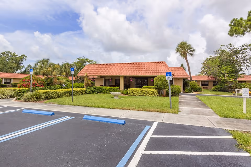Exterior view of a single-story building with a red tiled roof surrounded by green bushes and palm trees. There are several handicap parking spaces in the foreground with blue and white markings. The sky is partly cloudy.