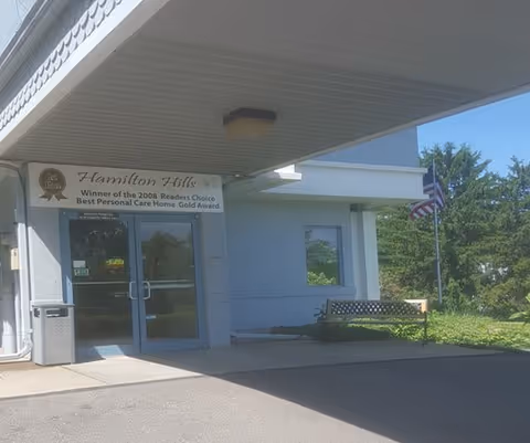 Entrance of Hamilton Hills senior care facility with glass doors, sign, bench, and flag.