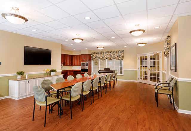 A spacious dining room with a long wooden table surrounded by green cushioned chairs. The room features wood flooring, beige and green walls, a mounted flat-screen TV, and a kitchen area with wooden cabinets and appliances in the background. There are windows with floral valances and a set of double glass doors.