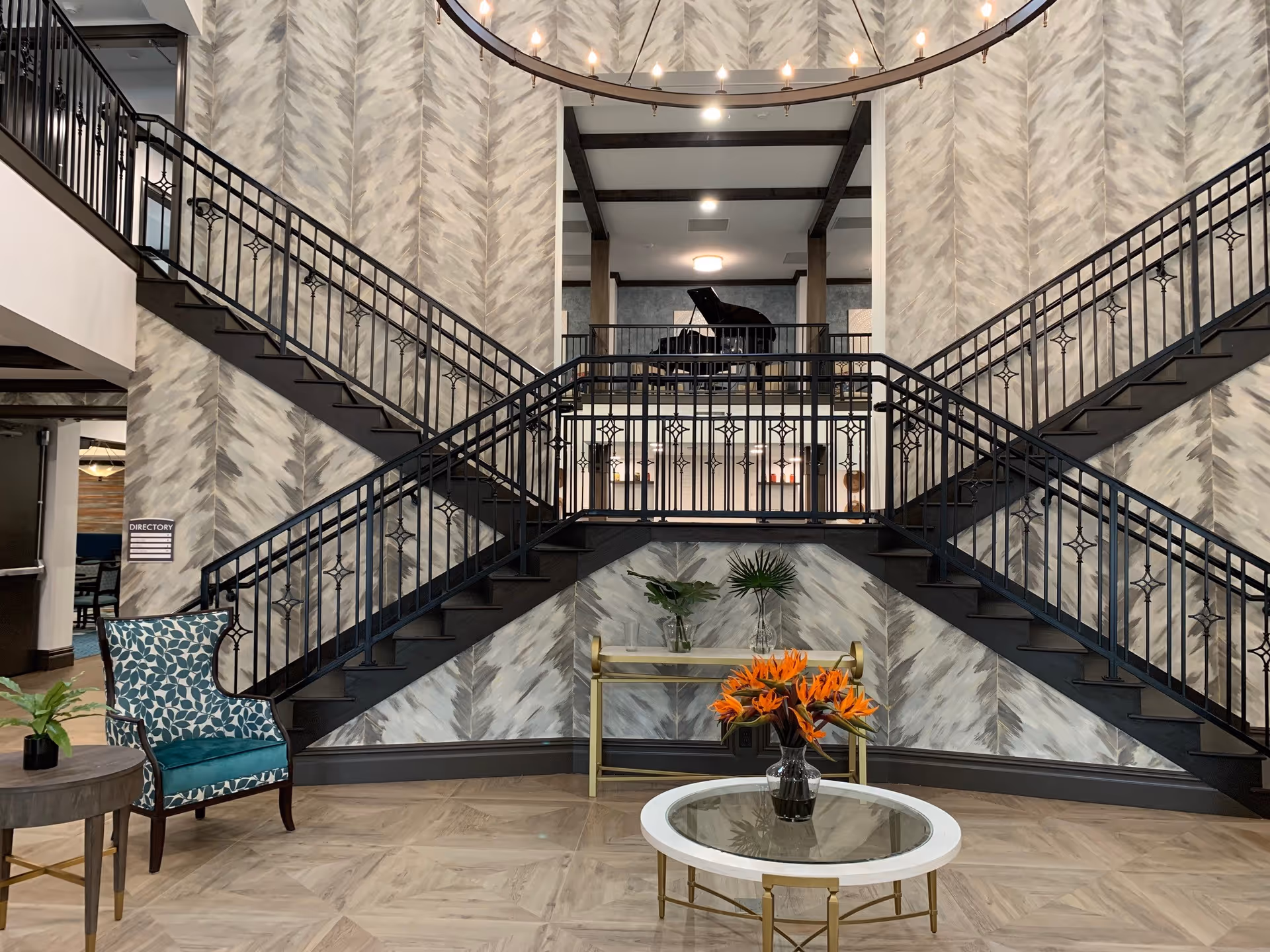 Spacious lobby with a symmetrical double staircase, chandelier, seating, floral arrangements and a grand piano on the mezzanine.