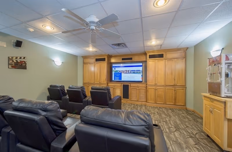 A cozy media room with several black leather recliners arranged in rows facing a large flat-screen TV mounted on a wooden entertainment center. The room has a ceiling fan, recessed lighting, light green walls, and a popcorn machine on a wooden cabinet to the right.