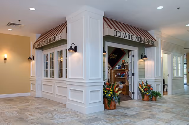 Interior view of a corner general store named 'DeLand General Store' inside a senior living facility. The store has white paneled walls with black wall-mounted lights and a beige awning with the store name. There are flower arrangements in pots placed near the entrance. The floor is tiled with a light stone pattern, and the surrounding area appears to be a hallway with additional rooms visible.