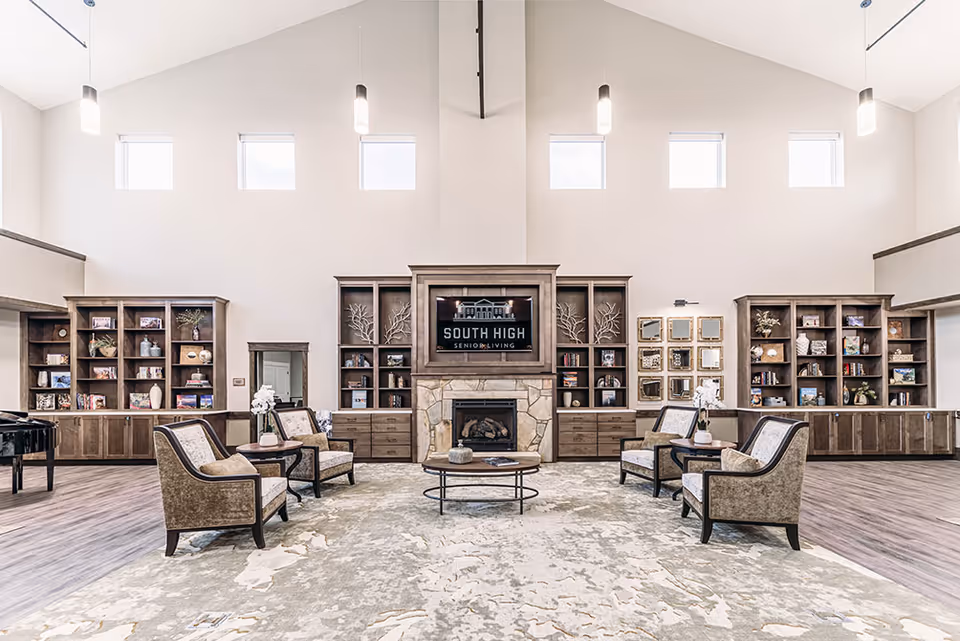 A spacious and well-lit senior living facility common area with high ceilings and multiple windows. The room features a central stone fireplace with a TV above it displaying 'South High Senior Living'. Surrounding the fireplace are built-in wooden bookshelves filled with decorative items and books. Four upholstered armchairs are arranged around a round coffee table on a large patterned rug. Pendant lights hang from the ceiling, and a grand piano is visible on the left side of the room.