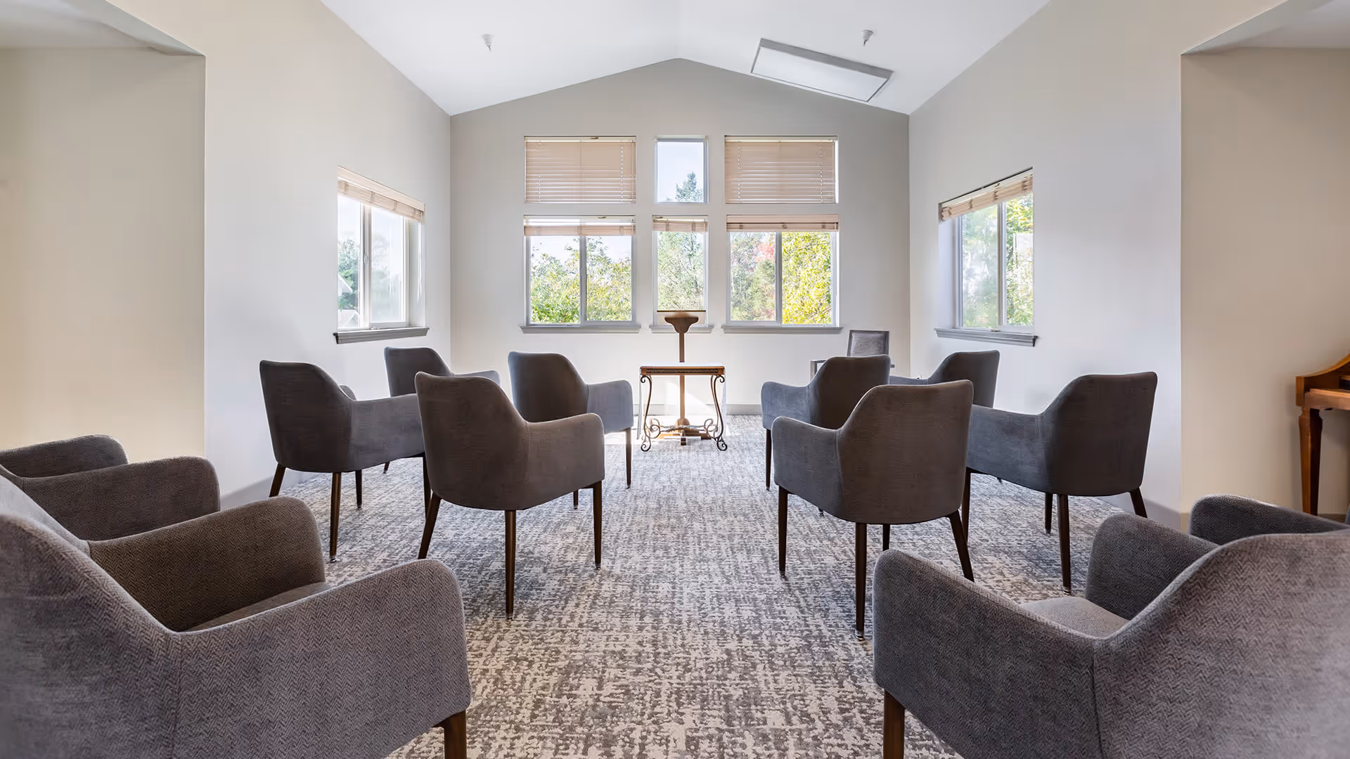 A bright room with large windows letting in natural light, arranged with multiple gray upholstered armchairs in a semi-circle facing a small wooden podium or lectern. The room has light-colored walls and a patterned carpet floor.