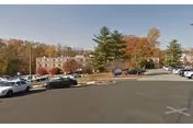 Parking lot and driveway leading to a brick multi-story building with parked cars and autumn trees.