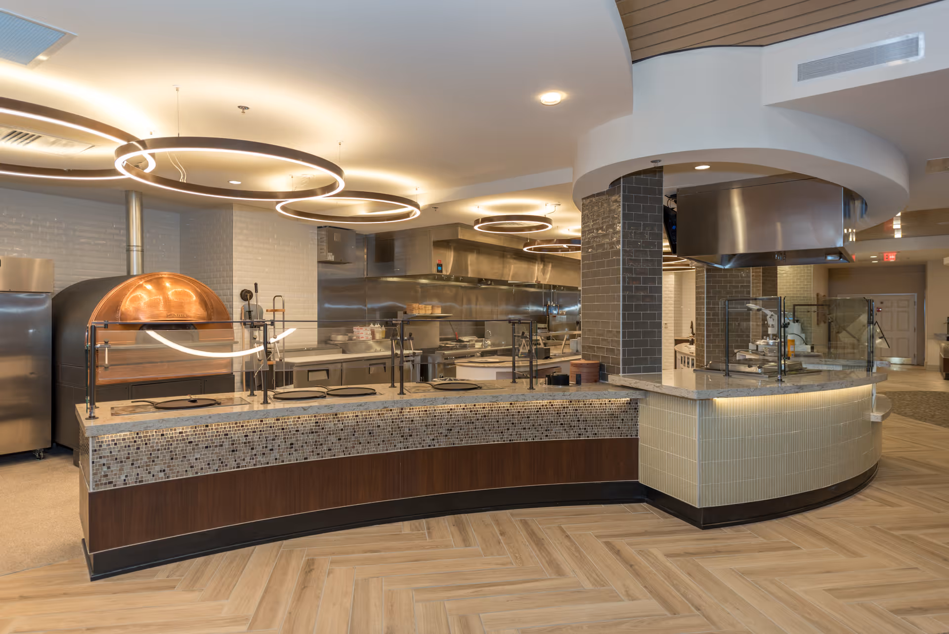 Modern open commercial kitchen and serving counter with a pizza oven, glass sneeze guards, and circular ceiling lights.