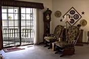 A cozy living room area with two cushioned rocking chairs facing a large sliding glass door that opens to a balcony. The room features a grandfather clock, decorative wall plates, and star-shaped wall decor. Light filters in through the glass door, illuminating the carpeted floor.