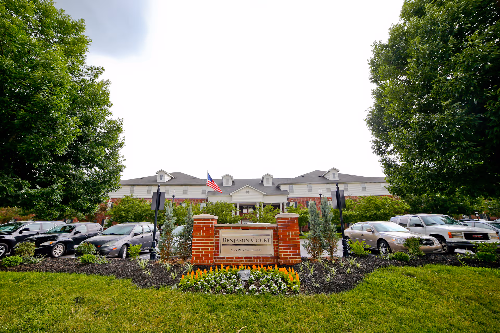 Front exterior view of Benjamin Court, a senior living facility, showing a brick sign with the facility name, an American flag, parked cars, and trees on either side.