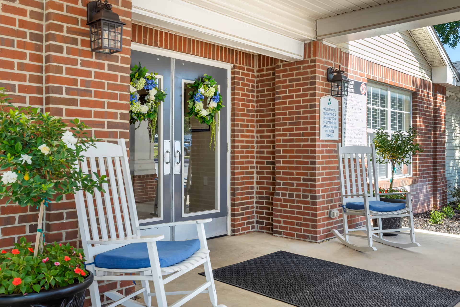 Entrance of a brick building with two white rocking chairs with blue cushions on either side of double glass doors decorated with floral wreaths. There are potted plants with flowers near the chairs and a black outdoor mat in front of the doors.