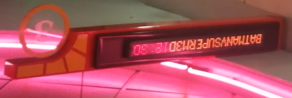 A digital sign displaying the text 'BATHRANSUFERM30 12:30' mounted on a wall with pink neon lighting around it.