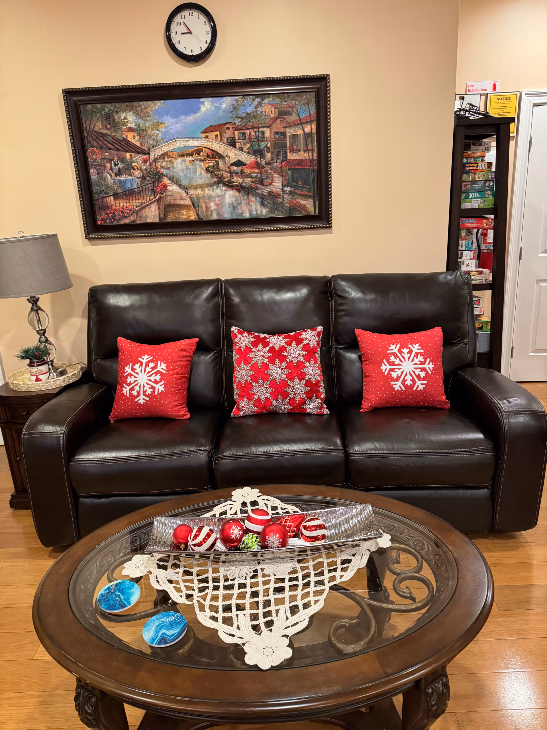 A cozy living room area with a dark brown leather sofa adorned with three red pillows featuring white snowflake designs. In front of the sofa is a wooden coffee table with a glass top, decorated with a white lace doily and a silver tray holding red and white Christmas ornaments. On the left side, there is a small wooden side table with a lamp and a small Christmas decoration. Above the sofa hangs a framed painting of a canal scene with a bridge and buildings. A clock is mounted on the wall above the painting. To the right, there is a bookshelf filled with board games and a white door.