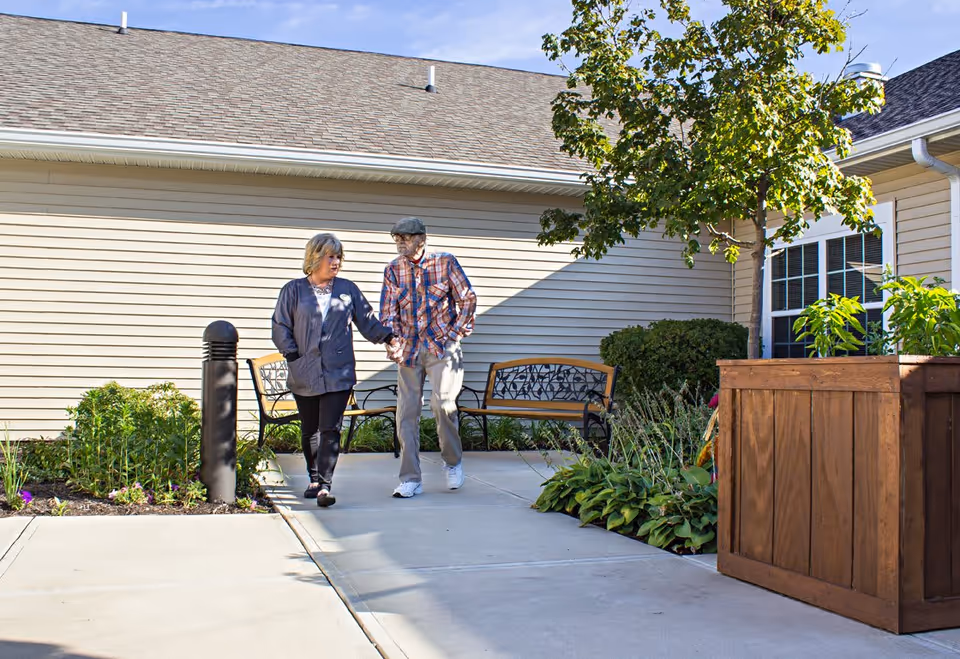 An elderly couple walking hand in hand on a paved pathway outside a senior living facility. They are surrounded by greenery, including bushes, plants, and a tree. There are benches along the side near the building with beige siding and a shingled roof under a clear blue sky.