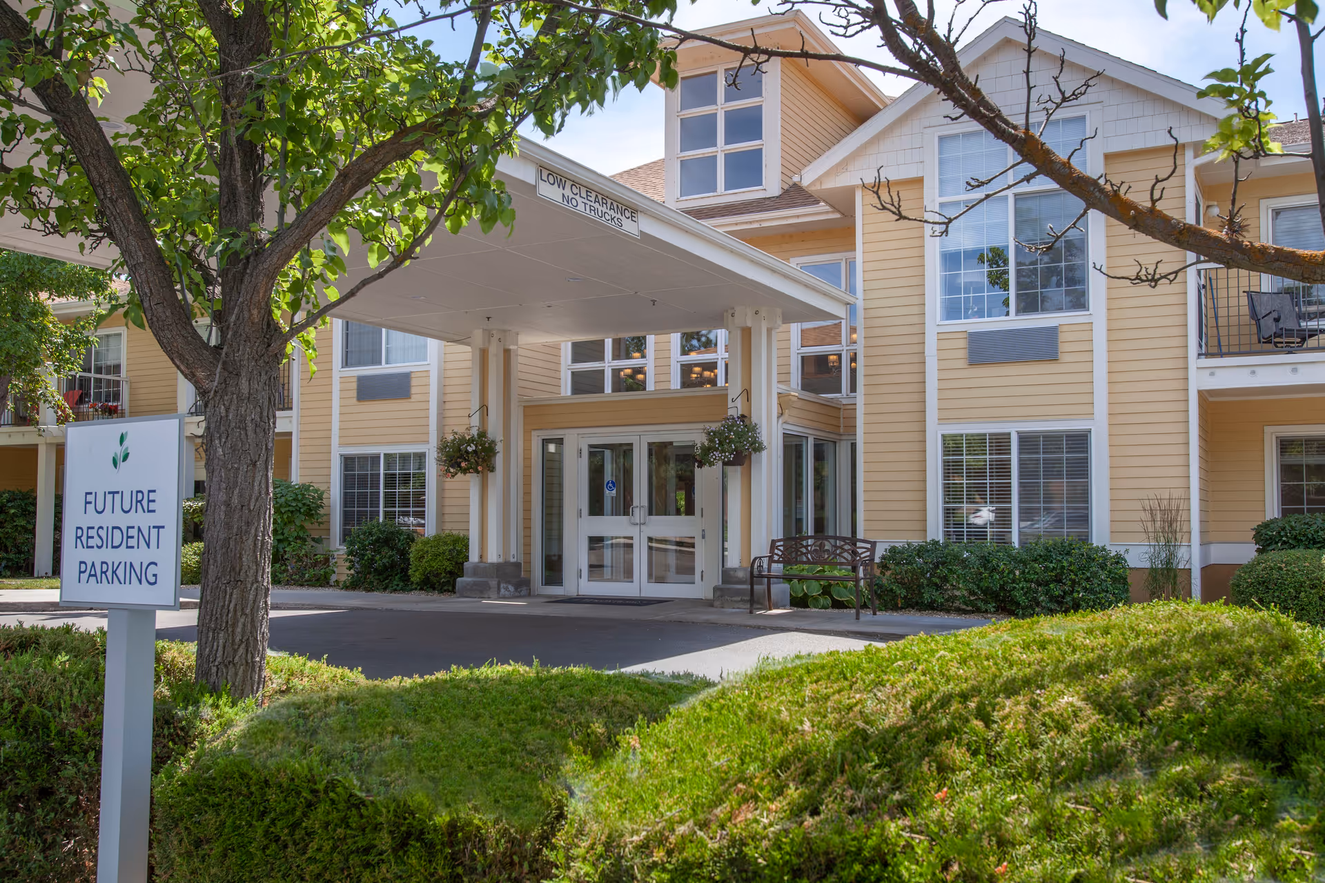 Exterior view of a senior living facility entrance with a covered drop-off area. The building is light yellow with white trim and multiple windows. There is a sign in the foreground that reads 'Future Resident Parking' next to a tree and green bushes. A bench is placed near the entrance doors.