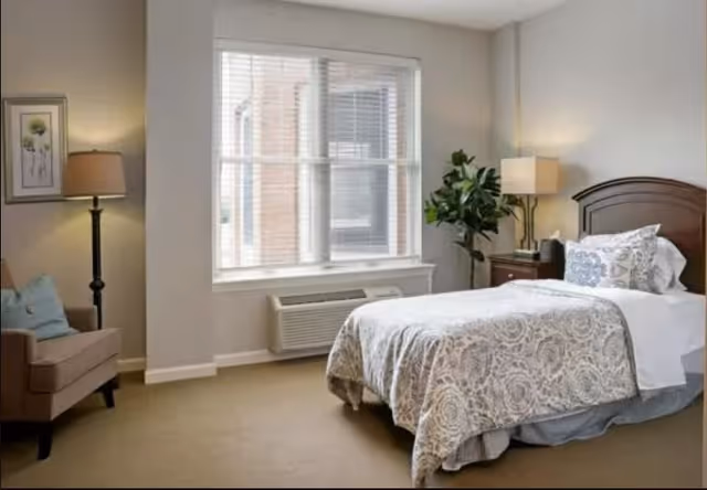 A neatly arranged bedroom with a single bed covered in a patterned bedspread and pillows. There is a wooden headboard, a nightstand with a lamp and a plant, a window with blinds, a beige armchair with a cushion, and a floor lamp next to the armchair. The walls are painted light gray and the floor is carpeted.