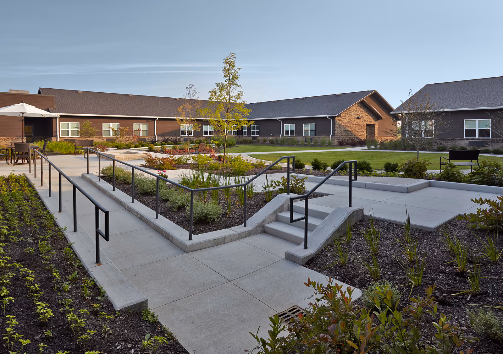 Well-maintained courtyard with paved walkways, ramps and railings, landscaping and seating in front of a single-story brick and siding senior living building.