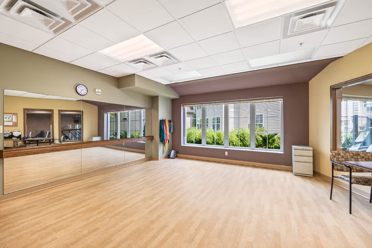 A spacious exercise or activity room with light wood flooring, large windows letting in natural light, a wall-length mirror, and some exercise bands hanging on the wall. There is a clock above the mirror and a chair with a patterned cushion near a glass door on the right side.