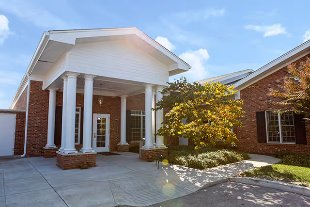 Entrance of a brick building with white columns supporting a covered porch, surrounded by greenery and a clear blue sky.