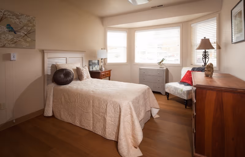 A cozy bedroom with a single bed covered in a beige quilt, a round dark brown pillow, and two beige pillows. Next to the bed is a wooden nightstand with a lamp and a framed picture. The room has a bay window with white blinds, a gray chest of drawers, a floral upholstered chair with a red pillow, and a wooden dresser with a lamp and decorative items. The walls are light beige, and there is a bird-themed painting on the wall.