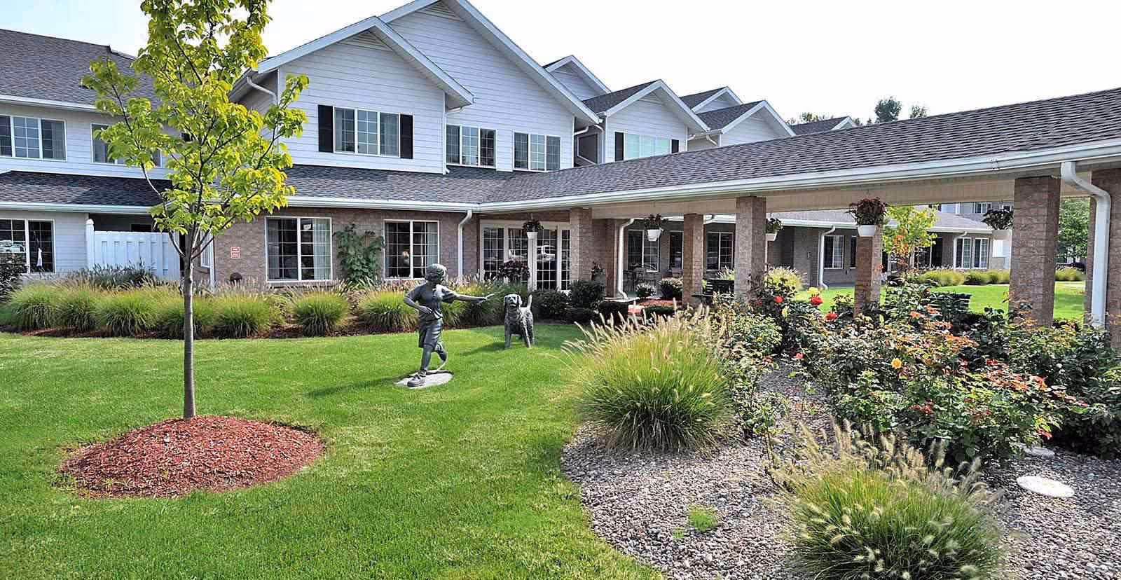 Outdoor garden area of a senior living facility with a green lawn, a young tree, flower beds, and a covered walkway supported by brick columns. There are two statues on the lawn, one of a child pointing and another of a dog. The building in the background has white siding and multiple windows.