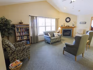 A cozy living room with a vaulted ceiling, featuring a floral patterned armchair and loveseat, two gray armchairs, a wooden bookshelf filled with books, a basket with magazines, a fireplace with a clock above it, and a large window with curtains letting in natural light.