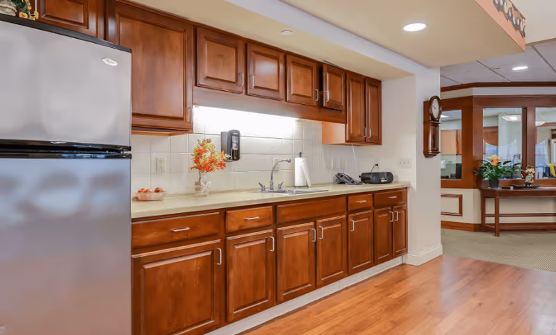Interior view of a kitchen area with wooden cabinets, a stainless steel refrigerator, a countertop with a sink, a paper towel holder, a phone, and a small flower arrangement. The floor is wooden and the kitchen opens into a carpeted area with a table and plants.