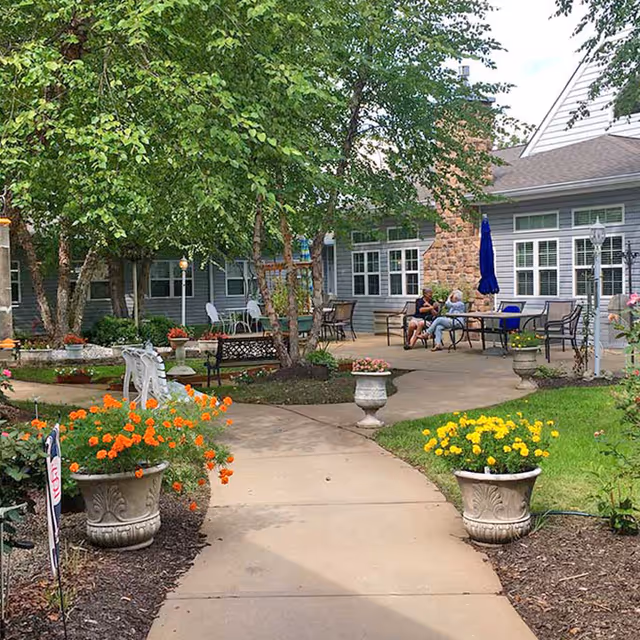 Outdoor courtyard area at Cape Albeon Assisted Living with a paved walkway, flower pots with orange and yellow flowers, benches, trees, and two people sitting and talking at a table near the building.