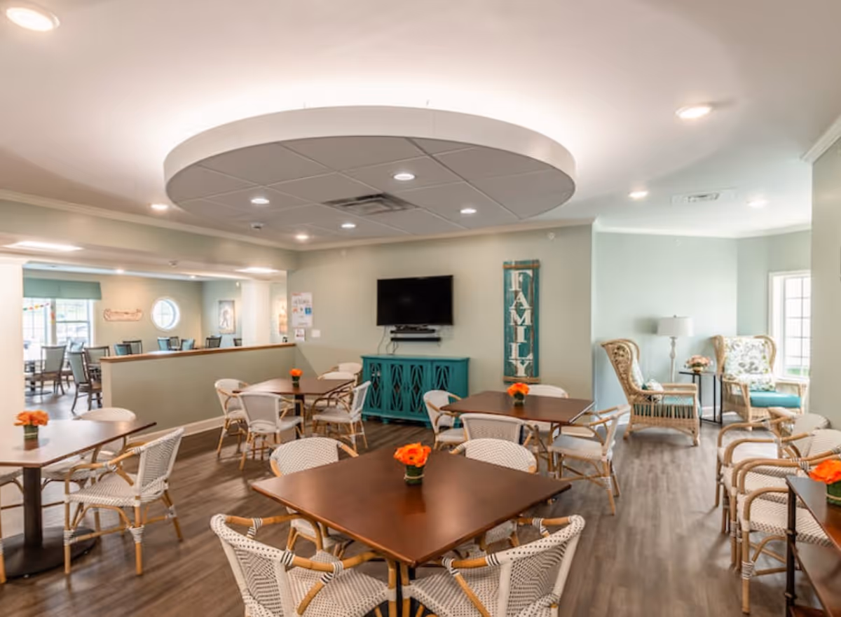 A bright and inviting common area with multiple tables and chairs arranged for socializing or dining. The room features a flat-screen TV mounted on the wall above a teal cabinet, a decorative sign that reads 'FAMILY,' and a cozy seating area with wicker chairs and a floor lamp near large windows letting in natural light. The ceiling has a circular recessed design with built-in lighting.