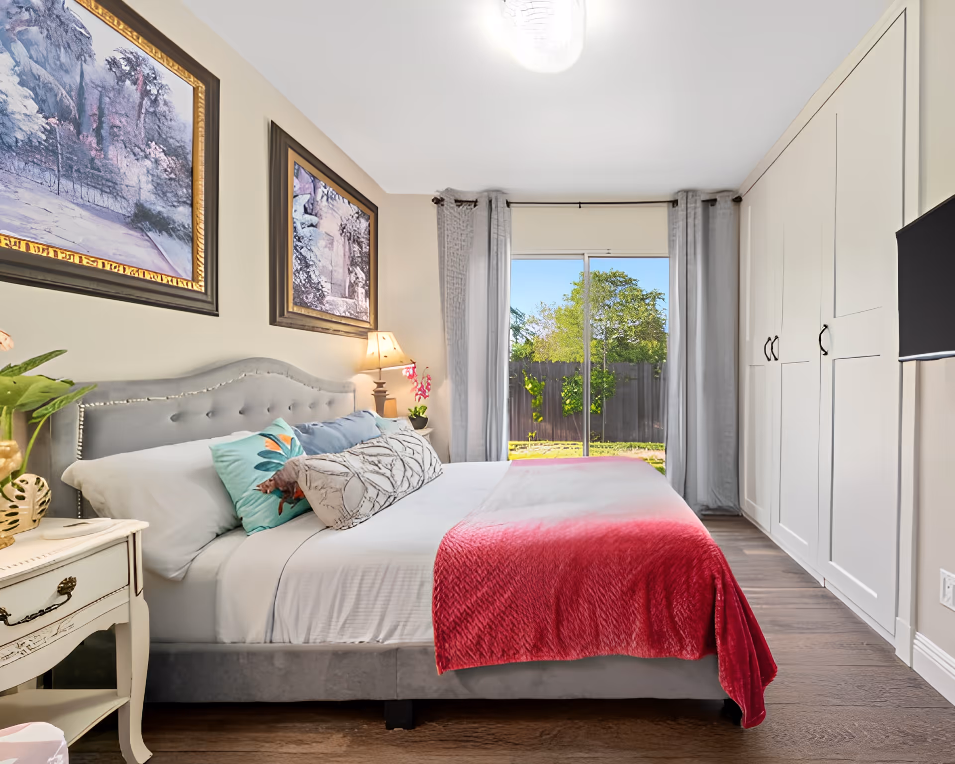 Bright, tastefully decorated bedroom with a bed, bedside tables, artwork, built-in wardrobes, and a sliding glass door to a yard.