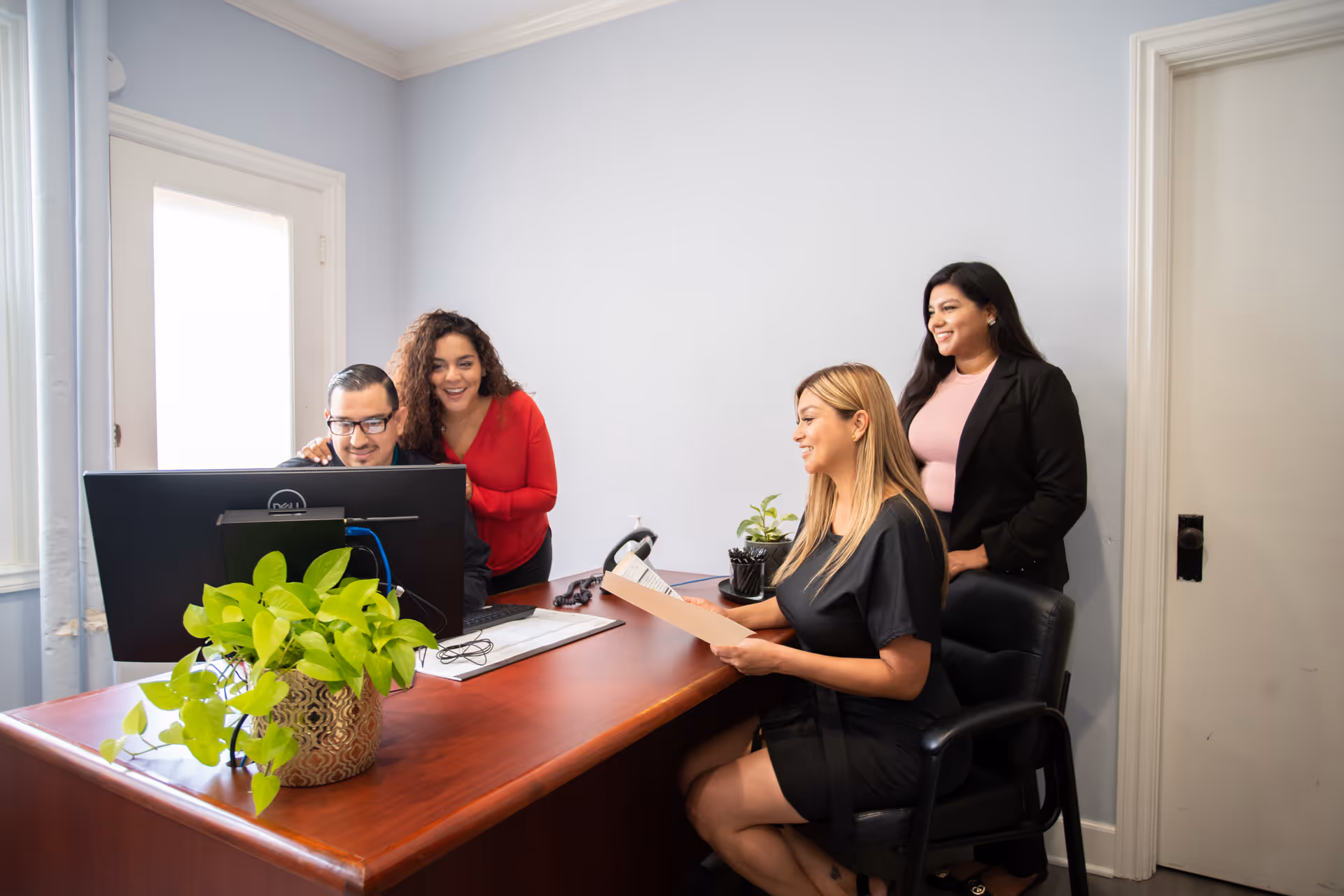 Four people in an office setting. One man is seated at a desk working on a computer while three women stand or sit around him, looking at the screen and a document. The office has light-colored walls, a wooden desk with a potted plant, and a door in the background.