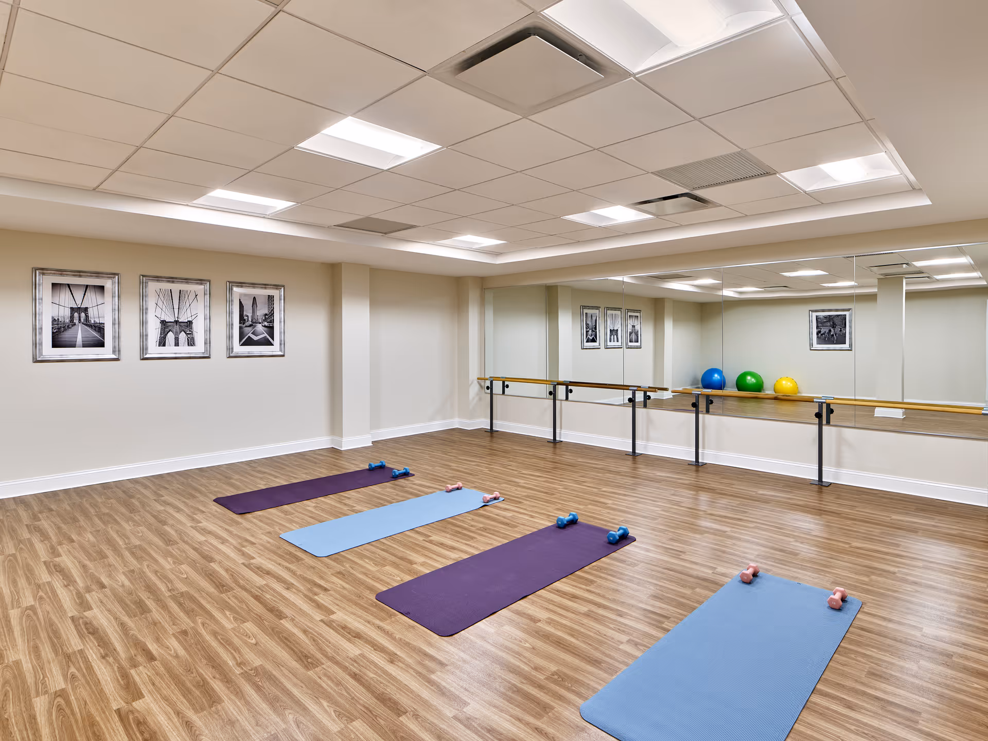 A spacious exercise room with wooden flooring and a mirrored wall. Four yoga mats are laid out on the floor, each paired with a set of small dumbbells. The room has a ballet barre along the mirrored wall and three framed black and white photos on the opposite wall. Three colorful exercise balls are placed on the floor near the mirror.