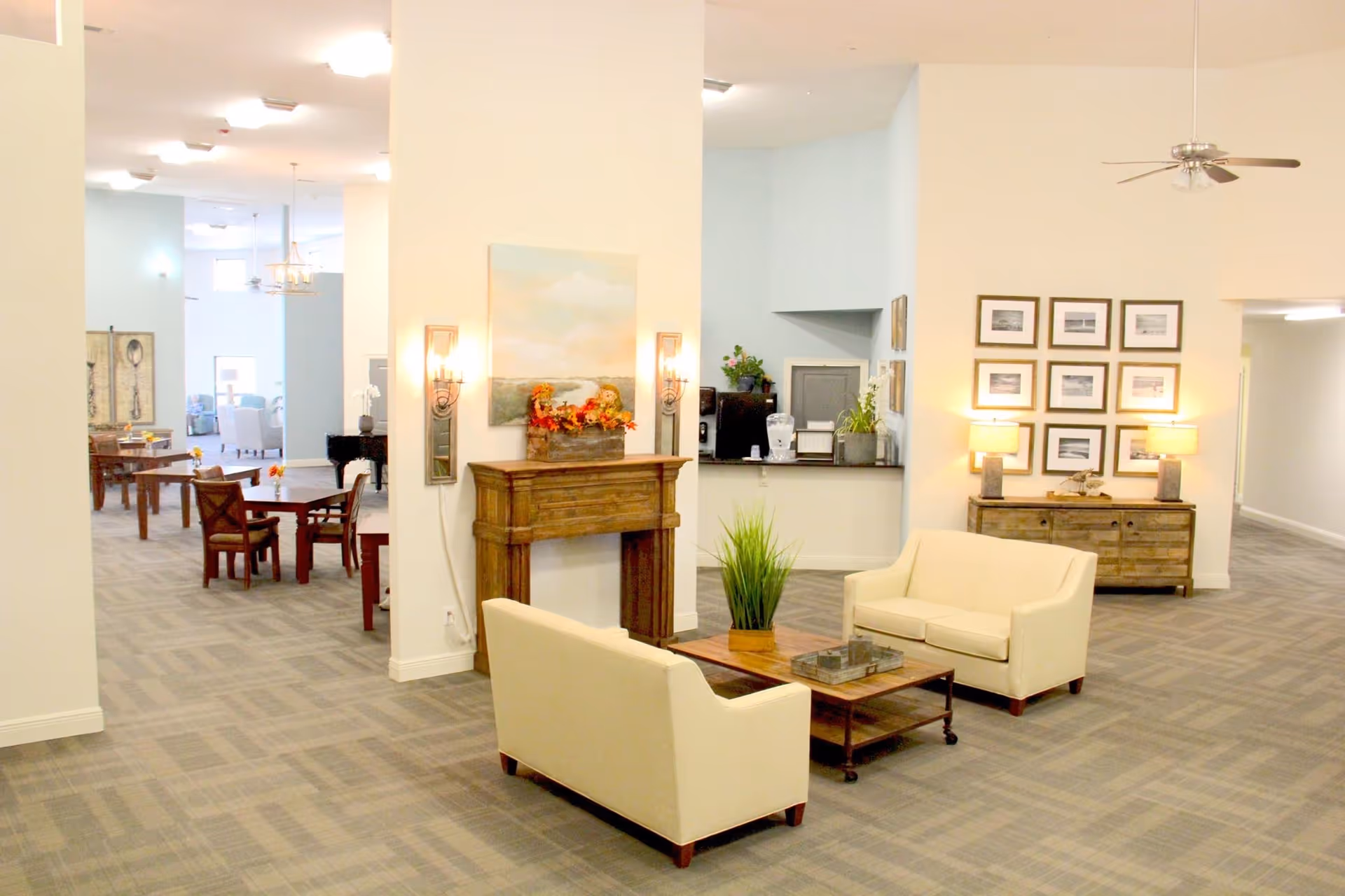 A spacious and well-lit common area in a senior living facility featuring two cream-colored sofas facing each other with a wooden coffee table in between. The room has a decorative wooden mantel with autumn-themed decor, a wall painting, and wall sconces. In the background, there is a dining area with tables and chairs, a piano, and a sideboard with framed pictures and lamps. The flooring is carpeted with a subtle pattern, and the walls are painted in light colors.