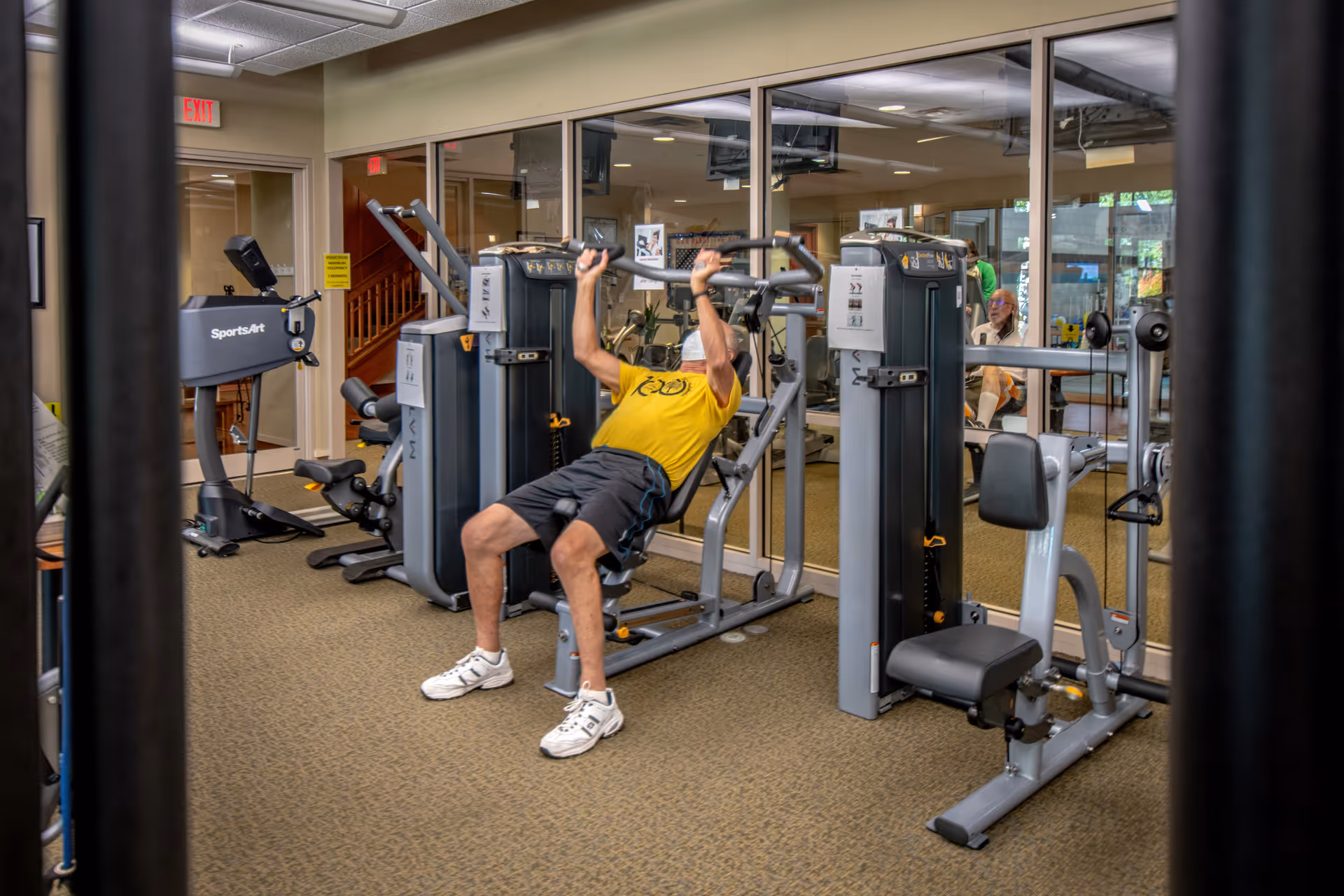 A man in a yellow shirt and black shorts is exercising on a weight machine in a fitness room. The room has various exercise equipment including a treadmill and other weight machines. There are large mirrors on the wall reflecting the room and other people in the background.