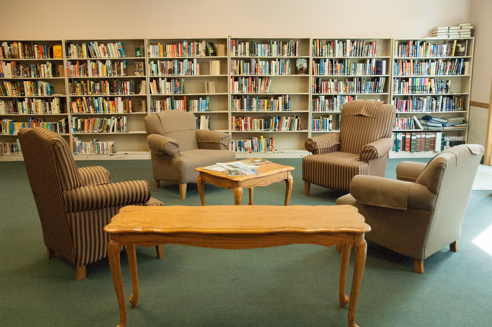 A cozy library or reading area with four upholstered armchairs arranged around a small wooden coffee table. Behind the chairs, there are tall bookshelves filled with books. The room has a green carpet and a wooden table in the foreground.