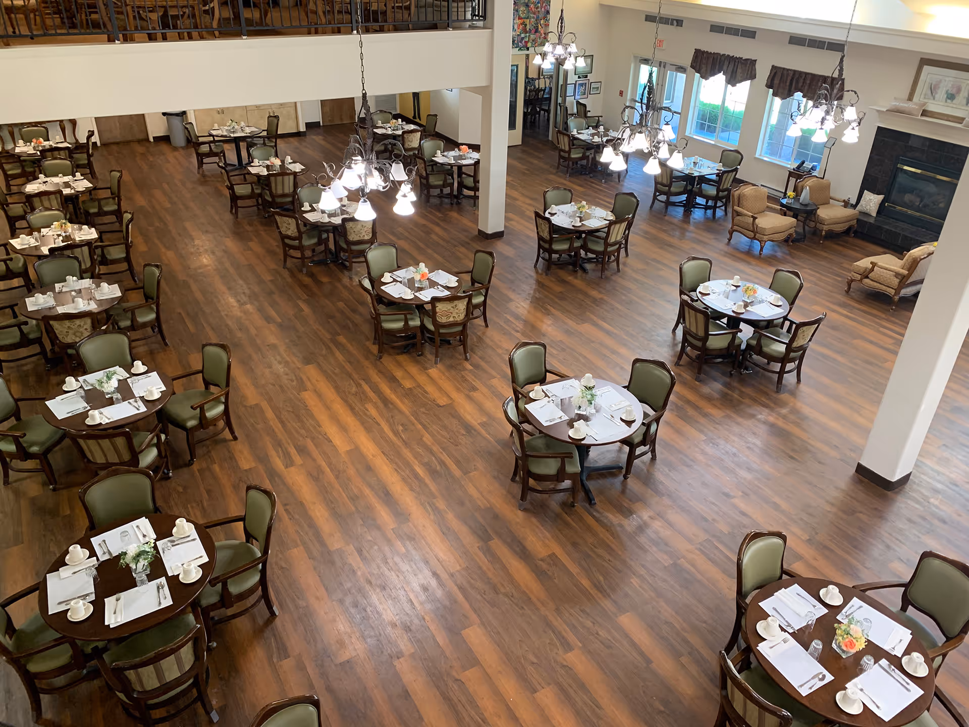 A spacious dining room with multiple round tables set with white napkins, cups, and silverware. The room has wooden flooring, green cushioned chairs, chandeliers hanging from the ceiling, and a seating area with armchairs near a fireplace and large windows.